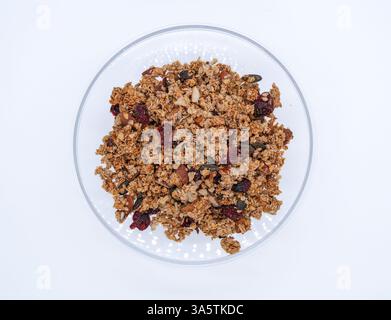 Un bol en verre transparent rempli de granola, avec un mélange d'avoine, de noix et de fruits secs, y compris des canneberges et des graines de citrouille, sur un fond blanc Banque D'Images