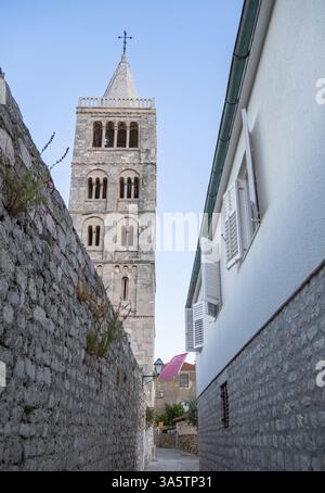 Rab, Croatie - 10 août 2024 : une ruelle étroite entre deux bâtiments, avec un grand clocher en pierre en arrière-plan. La tour a une croix en Banque D'Images