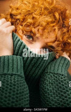 Femme avec des cheveux bouclés de gingembre couvrant son visage avec un pull en laine verte Banque D'Images