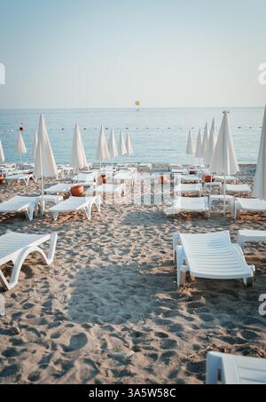 Une scène de plage tôt le matin avec des chaises longues vides et des parasols fermés disposés soigneusement sur le sable. La mer calme et le parasailer lointain ajoutent à la Banque D'Images