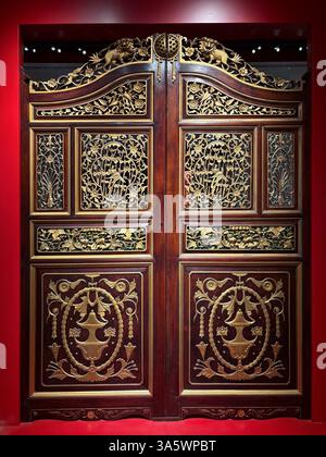 Une paire de portes en bois brun foncé finement sculptées avec des détails dorés, serties contre un mur rouge à Singapour. Banque D'Images