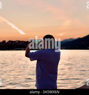 Une personne capture le coucher de soleil serein sur un lac tranquille à l'aide d'un smartphone, avec des montagnes et un ciel orange comme toile de fond, exprimant la paix Banque D'Images