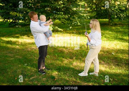 Les parents et l'enfant passent les week-ends d'été ensemble dans le parc. Maman souffle des bulles, papa tient mignon enfant dans ses bras, ils rient et s'amusent ensemble. Inter Banque D'Images