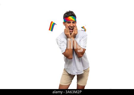 Jeune homme latin portant un bandeau arc-en-ciel vibrant et tenant des drapeaux colorés, exprimant la surprise et la joie tout en célébrant la fierté gay dans un s joyeux Banque D'Images