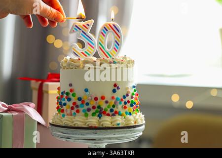 30e anniversaire. Femme allumant des bougies en forme de nombre sur le gâteau contre fond flou avec des lumières, gros plan. Espace pour le texte Banque D'Images