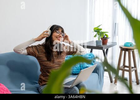 Heureuse freelance travaillant depuis son salon, profitant de la musique avec des écouteurs tout en utilisant un ordinateur portable Banque D'Images