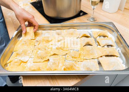 Un plateau de raviolis fraîchement préparés saupoudrés de farine, prêt à cuire. Une main ramasse une pièce, mettant en valeur la préparation artisanale des pâtes. Banque D'Images