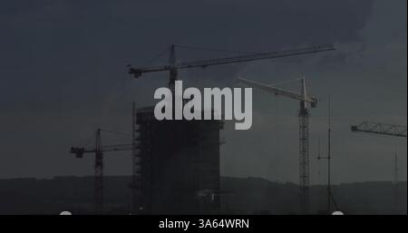 Grues de construction surplombant un immeuble de grande hauteur sous un ciel sombre et nuageux Banque D'Images