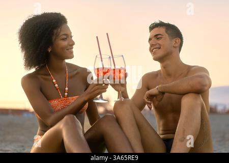 Jeune couple interracial toast avec Aperol Spritz sur la plage au coucher du soleil. Un moment romantique et joyeux de vacances d'été, célébrant l'amour, Happin Banque D'Images