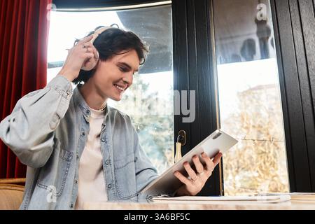 Un jeune homme sourit dans un café, profitant de la musique avec des écouteurs tout en utilisant sa tablette. Banque D'Images