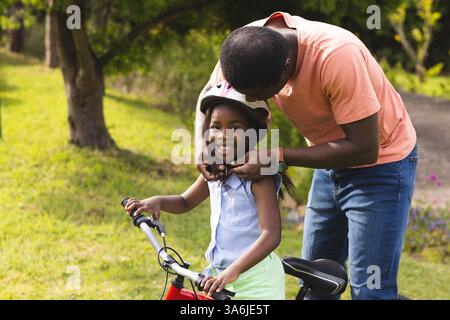 Père aidant sa fille avec un casque alors qu'elle sourit sur le vélo à l'extérieur Banque D'Images