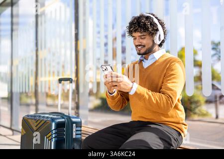 Un jeune homme avec des écouteurs attend à un arrêt de bus, regardant son smartphone avec sa valise à côté de lui par une belle journée. Banque D'Images