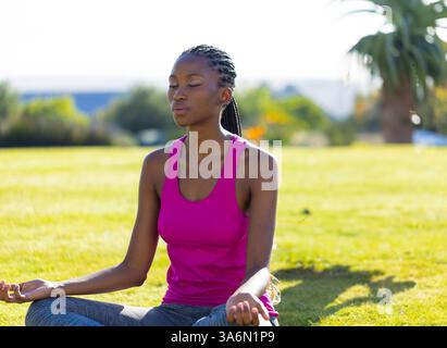Femme afro-américaine méditant sur l'herbe dans l'arrière-cour, profitant d'un moment paisible Banque D'Images
