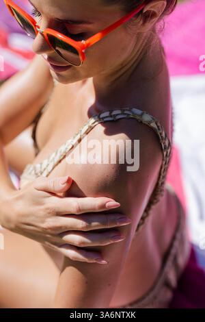 Femme dans des lunettes de soleil appliquant de la crème solaire par piscine, profitant de la journée ensoleillée à l'extérieur Banque D'Images