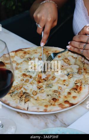 Pizza blanche à quatre fromages coupée par des mains féminines sur un contexte de restaurant, vin rouge sur le côté. Les vibrations du déjeuner Banque D'Images
