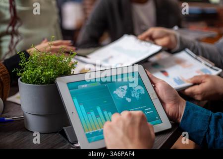 Une personne caucasienne tient une tablette affichant l'analyse de marketing de l'entreprise avec une plante verte à proximité sur le bureau. Gros plan sur l'écran de l'appareil montrant des graphiques de données, visualisés par un individu lors d'une réunion. Banque D'Images
