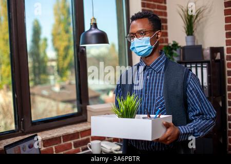 Employé afro-américain portant un masque facial et portant une boîte de matériel de travail, se préparant à quitter le bureau. Dans le lieu de travail de mur de briques, le gérant avec des lunettes se tient près de la fenêtre tenant ses affaires. Banque D'Images