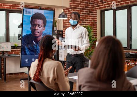 Homme noir dans le masque facial gestes à l'écran avec videocall actif du client afro-américain, fournissant des mises à jour de projet. L'équipe s'engage dans une réunion virtuelle avec le gestionnaire à distance, analysant les indicateurs de l'entreprise. Banque D'Images