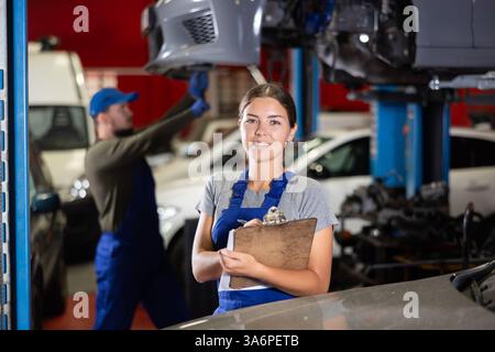 Travailleuse de service automobile tient des registres du travail effectué par les mécaniciens automobiles dans la station-service automobile - prend des notes sur papier Banque D'Images