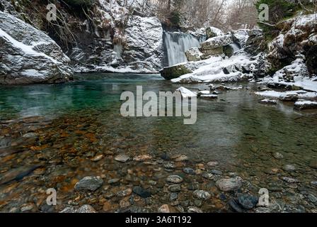 La neige recouvre le terrain rocheux tandis qu'une cascade se déverse dans une piscine claire, créant un paysage hivernal paisible dans la forêt. Banque D'Images