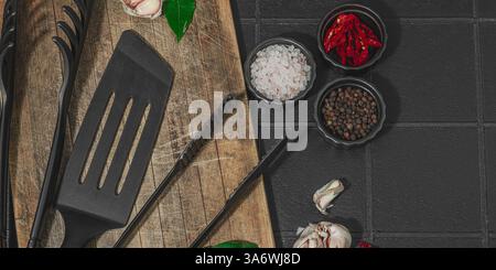 Concept de saison barbecue. Pinces à griller, spatule, set d'épices, gros sel, ail et herbes fraîches. Préparation des aliments grillés, fond de tuiles noires, plat Banque D'Images