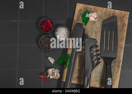 Concept de saison barbecue. Pinces à griller, spatule, set d'épices, gros sel, ail et herbes fraîches. Préparation des aliments grillés, fond de tuiles noires, plat Banque D'Images