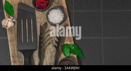 Concept de saison barbecue. Pinces à griller, spatule, set d'épices, gros sel, ail et herbes fraîches. Préparation des aliments grillés, fond de tuiles noires, plat Banque D'Images