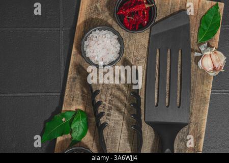 Concept de saison barbecue. Pinces à griller, spatule, set d'épices, gros sel, ail et herbes fraîches. Préparation des aliments grillés, fond de tuiles noires, plat Banque D'Images