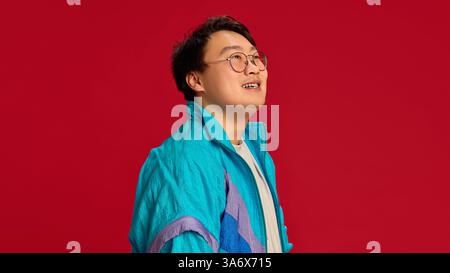 Vue latérale gros plan portrait d'un jeune homme asiatique portant des lunettes avec un sourire léger regardant loin sur fond rouge. Banque D'Images