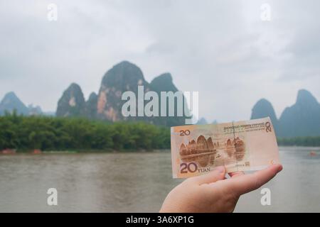 Une main tenant un billet chinois de 20 yuan correspondant au paysage karstique réel de la rivière Li à Guilin par une journée nuageuse dans le Guangxi, en Chine Banque D'Images