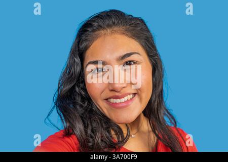 Portrait d'une jeune femme latine joyeuse avec de longs cheveux foncés, souriant sur un fond bleu vif Banque D'Images