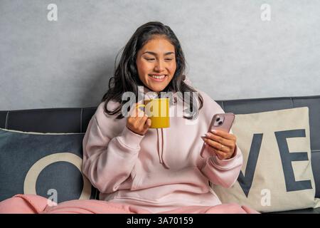 Heureuse jeune femme assise confortablement sur un canapé, profitant d'une tasse de café tout en naviguant sur son smartphone Banque D'Images