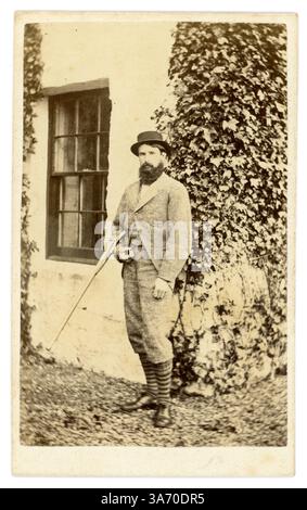 Carte de visite victorienne originale (carte de visite ou CDV) d'un jeune homme victorien barbu attrayant, gentleman victorien, appelé M. John Bateson, portant des vêtements de sport de pays de noblesse débarqué, posant pour une photographie devant sa maison, tenant un fusil de chasse. D'après une collection de la famille Arkwright, Hampton court Castle Hereford, photographié par Marcus Ward & Co 13 Donegall place, Belfast c1867 Banque D'Images