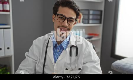 Beau jeune médecin masculin hispanique souriant dans la chambre d'hôpital moderne portant un manteau blanc et stéthoscope, mettant en valeur la pratique médicale professionnelle en cl Banque D'Images