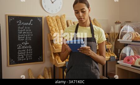 Femme travaillant dans une boulangerie vérifiant une tablette portant un tablier rayé, avec divers pains et pâtisseries affichés en arrière-plan Banque D'Images