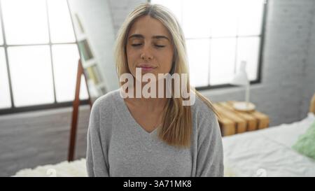 Jeune femme aux cheveux blonds est assise paisiblement avec les yeux fermés dans une chambre lumineuse, respirant le calme et la détente dans un cadre intérieur aéré et moderne. Banque D'Images