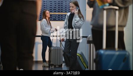 Terminal de l'aéroport bondé : femme utilisant un smartphone, attendant le vol dans le salon d'embarquement du Hub de la compagnie aérienne, vérifiant les informations à l'arrivée et au départ du tableau. Personnes diverses et personnel de l'aéroport marchant. Banque D'Images