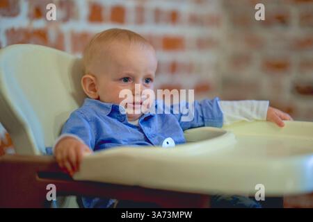 Un garçon de 1 an dans une chemise est assis dans une chaise haute dans un restaurant et sourit . Photo de haute qualité Banque D'Images
