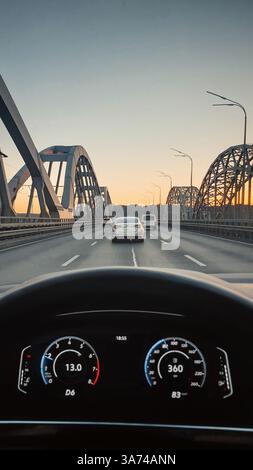Vue à la première personne de conduire une voiture sur un pont au coucher du soleil. Point de vue du conducteur : assis dans une voiture moderne avec tableau de bord numérique sur le pont autoroutier. Banque D'Images