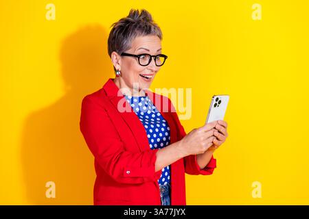 Femme d'âge moyen excitée avec les cheveux gris courts à la mode naviguant sur smartphone tout en portant une veste rouge sur fond jaune vif Banque D'Images