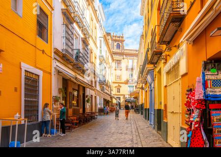 Les piétons passent devant les boutiques et les cafés sur les trottoirs à travers une rue étroite et colorée dans le quartier Barrio Santa Cruz de Séville en Espagne. Banque D'Images