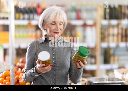 Shopping dans le supermarché de produits - acheteur fremale mature fait un choix lors de l'achat d'olives en conserve Banque D'Images