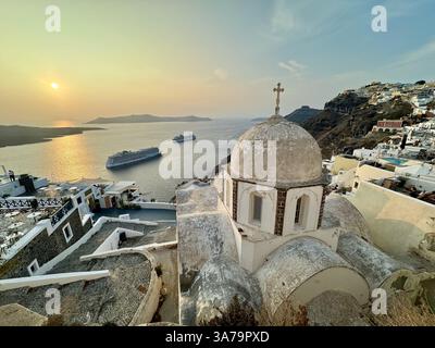 Une vue surplombant le dôme de l'église de Saint Jean le théologien à Santorin au coucher du soleil, avec deux grands bateaux de croisière à l'ancre dans la caldeira. Banque D'Images