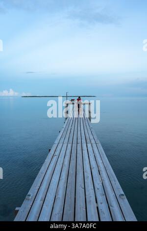 Une matinée paisible sur la plage de Maupiti, en Polynésie française, avec une jetée en bois s’étendant sur des eaux calmes, où une femme marche vers l’horizon au soleil Banque D'Images