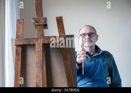 Un artiste masculin plus âgé, portant des lunettes et un tablier, peignant soigneusement sur une toile à l'aide d'un chevalet en bois dans une pièce bien éclairée. Banque D'Images