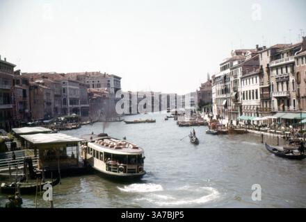Une vue pittoresque sur le Grand canal de Venise, prise en 1965, avec ses palais historiques, ses gondoles et ses bateaux-taxis. Le canal, bordé d'une élégante architecture vénitienne, serpente au cœur de la ville, servant de voie navigable principale. Des gondoliers naviguent habilement sur le canal, tandis que des vaporettos (bateaux-bus publics) transportent les passagers à travers ce paysage intemporel. Banque D'Images