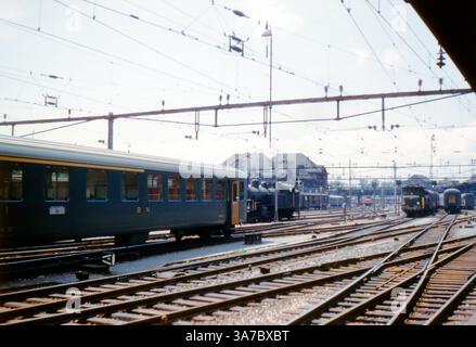 Une scène animée à la gare de Bâle en Suisse dans les années 1960, capturée sur film 35mm. La photo montre plusieurs voies avec des fils électriques au-dessus, un mélange de locomotives électriques et à vapeur, et des voitures de passagers. Ce hub ferroviaire dynamique reflète l'ère de la transition dans le transport ferroviaire européen de la vapeur à l'électricité. Banque D'Images
