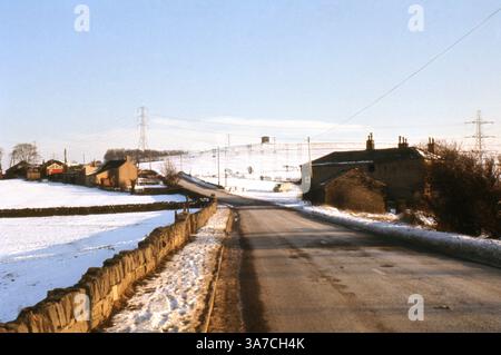 Une route de campagne s'étend à travers la banlieue enneigée de Dewsbury, West Yorkshire, en avril 1969. Des murs de pierre et des fermes traditionnelles bordent le bord de la route, avec des poteaux télégraphiques et des pylônes traçant la route vers des collines recouvertes de neige lointaines. Une tour ronde solitaire se dresse à l'horizon, ajoutant une qualité intemporelle à cette scène rurale hivernale. L'image capture le paysage de transition entre la ville et la campagne pendant un printemps britannique d'après-guerre. Banque D'Images