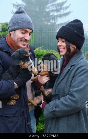 Jeune couple tenant leurs deux adorables teckshunds, souriant et profitant d'une journée brumeuse dans la nature Banque D'Images
