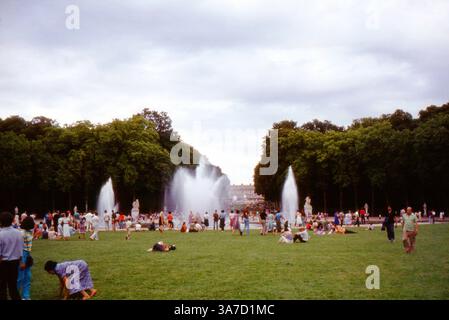 Les visiteurs se rassemblent autour des spectaculaires fontaines du bassin d’Apollon dans les jardins de Versailles, France. Ces scènes de fin d'été de 1984 montrent des foules appréciant la grandeur du jardin à la française, tandis que de puissants jets d'eau éclatent de la célèbre fontaine Apollon. Banque D'Images